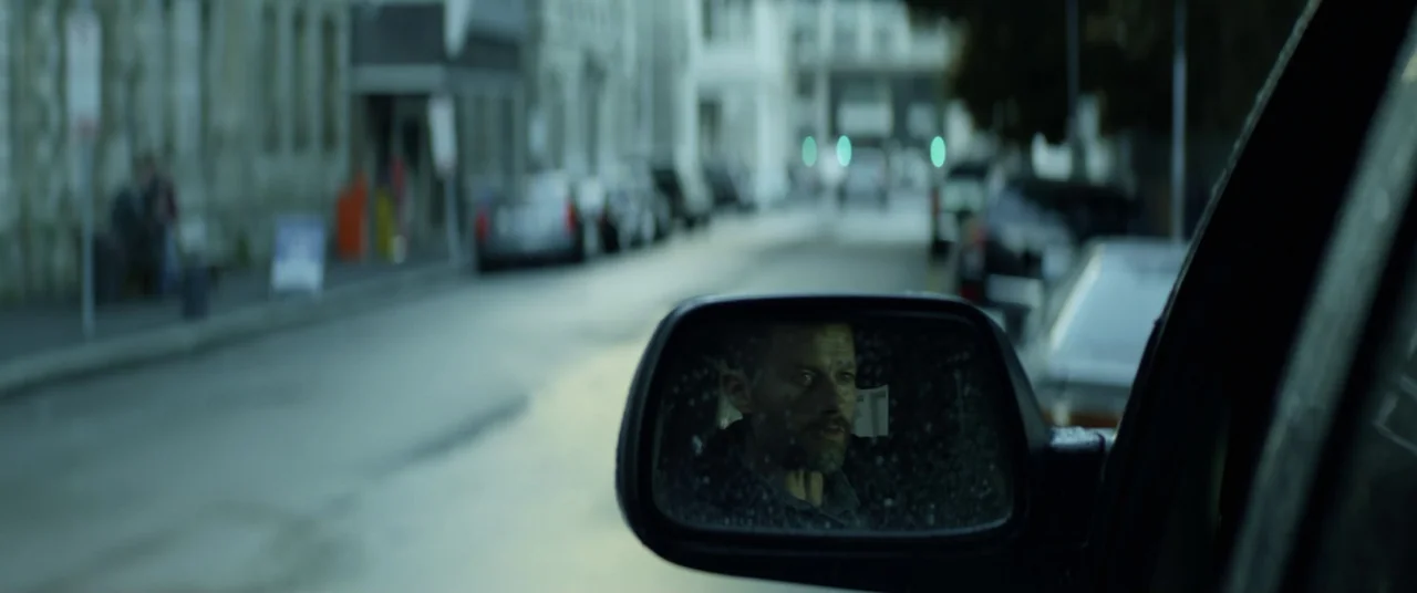 The Empty Man (2020) movie still: wide — A city street with parked cars is visible beyond a close-up side-view mirror. A man is visible inside the…