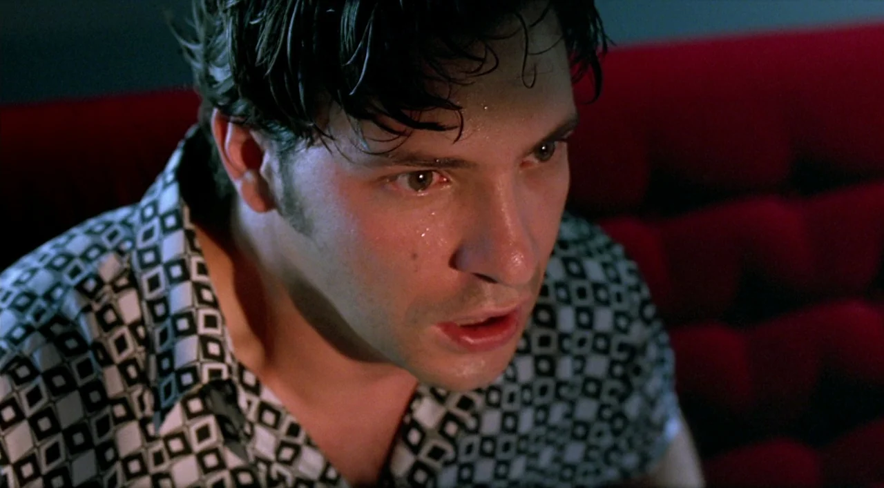 Kika (1993) movie still: closeup — A dark indoor space features a subject seated against a textured red backdrop. A man with perspiration on his…