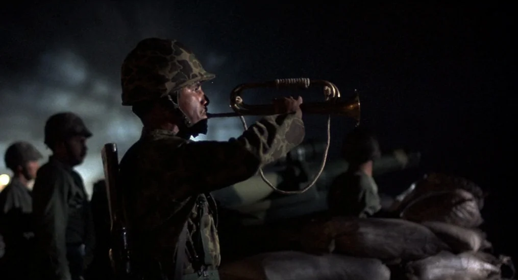 Salvador (1986) movie still: medium — A dark outdoor position with sandbags and soldiers standing in the background. One soldier in a camouflage…