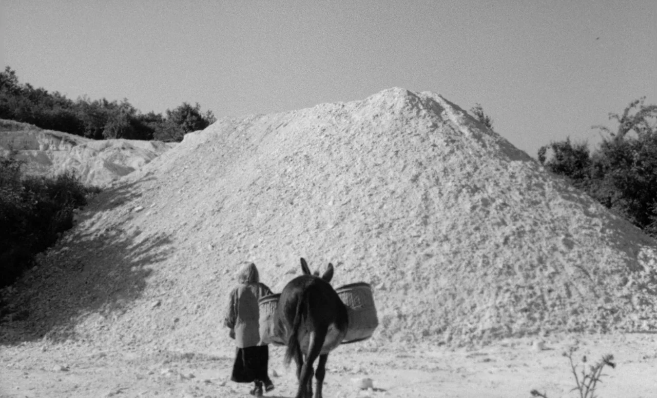 Goto, Island of Love (1969) movie still: wide — A large pile of white rock rises from a flat dusty clearing surrounded by vegetation. A person walks…