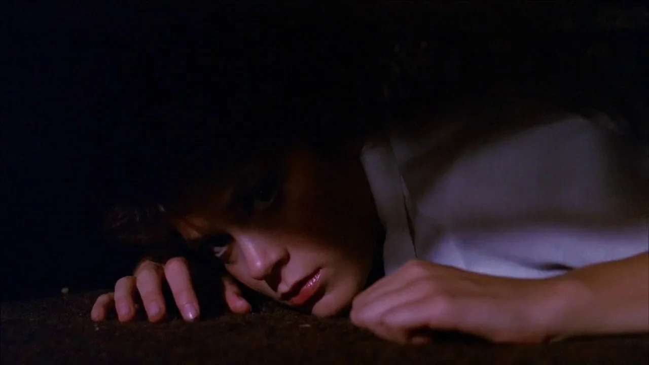 The Slumber Party Massacre (1982) movie still: closeup — A woman lies flat on a dark textured surface in deep shadow. She is positioned close to the…