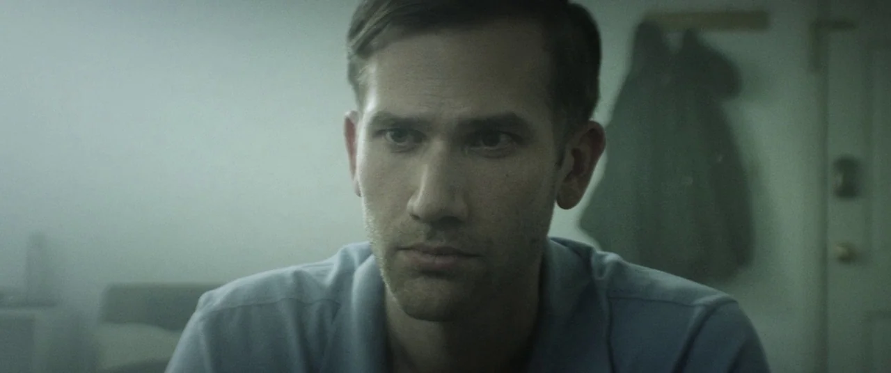 The Endless (2017) movie still: medium — A man stands in a dimly lit room with muted green tones and fog. He stares ahead while a coat hangs on a…