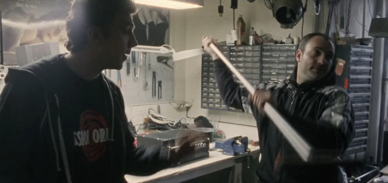 Pusher (1996) movie still: medium — A cluttered workshop contains a metal workbench, storage bins, and tools on a pegboard wall. One man holds a…