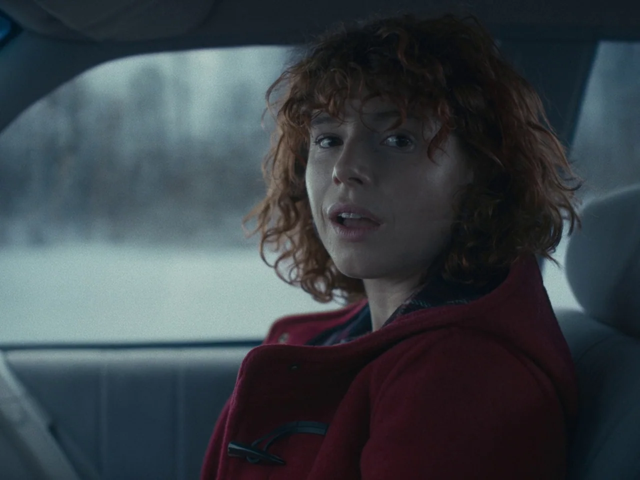 I’m Thinking of Ending Things (2020) movie still: medium — A woman sits in the driver seat of a car during a winter day. She is wearing a red coat…