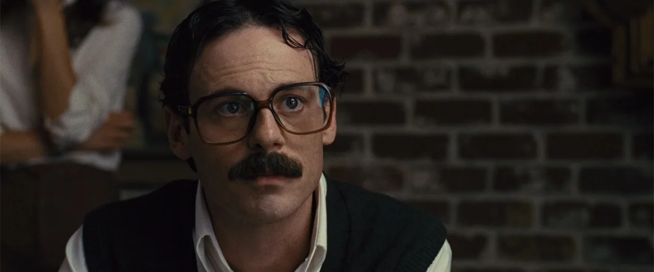 Argo (2012) movie still: closeup — A man stands before a blurred brick wall in an interior space. The subject stares forward with a blank expression…