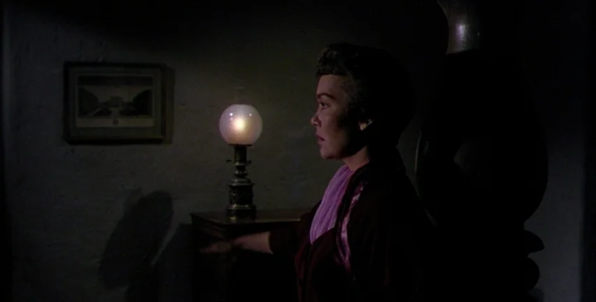 Magnificent Obsession (1954) movie still: medium — A dim room with a framed picture on the wall and an oil lamp. The woman stands in profile by a…
