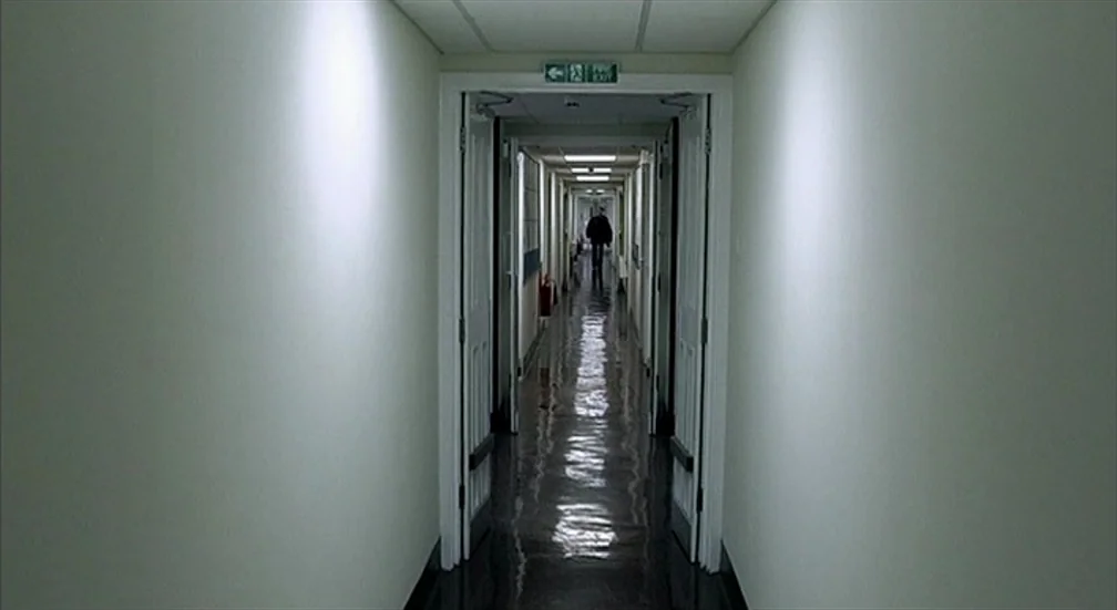 9 Songs (2004) movie still: wide — A long, symmetrical hospital corridor features off-white walls and highly reflective dark floors. A lone…