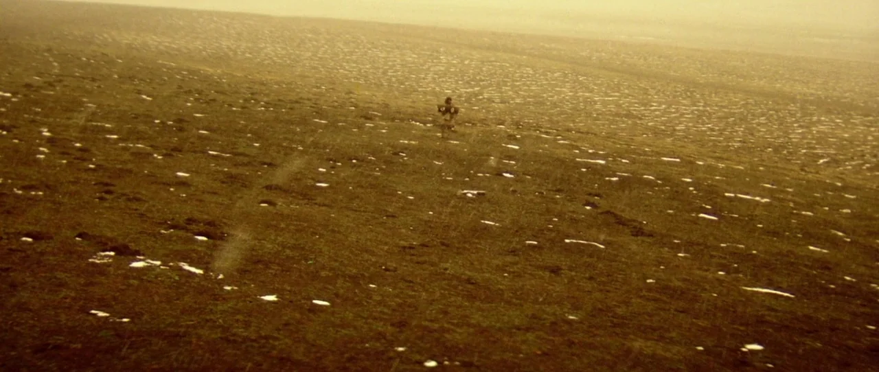 Horse Thief movie still: wide — A vast, open plain with patches of snow scattered across dark, barren ground. A lone person walks slowly across the…