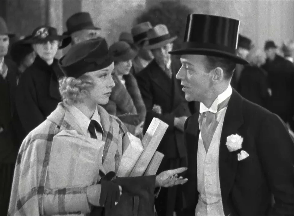Swing Time (1936) movie still: medium — A crowd of people in 1930s formal attire mills about an outdoor public space. A woman in a plaid coat speaks…