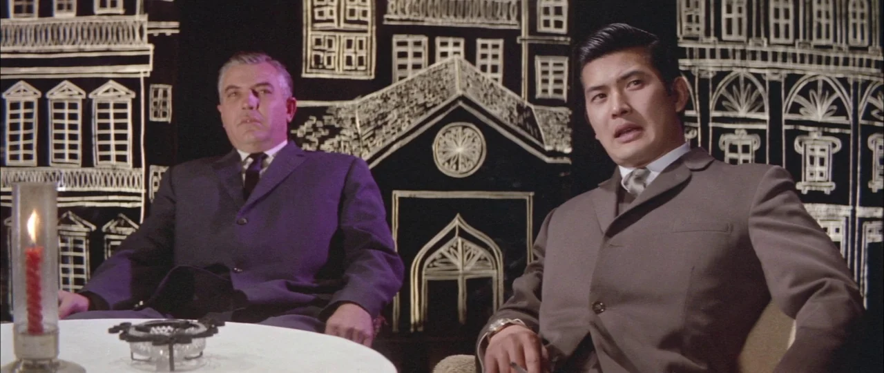 Black Tight Killers (1966) movie still: medium — A studio set displays a stylized black backdrop with white painted architectural line drawings. Two…