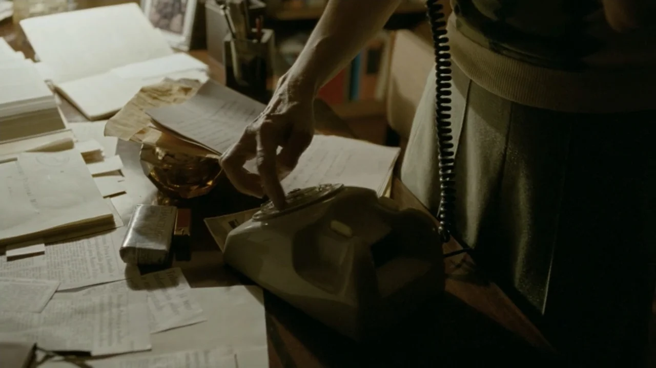 I'm Still Here (2024) movie still: medium — A cluttered wooden desk is covered in numerous stacked papers and a rotary telephone. A person wearing a…