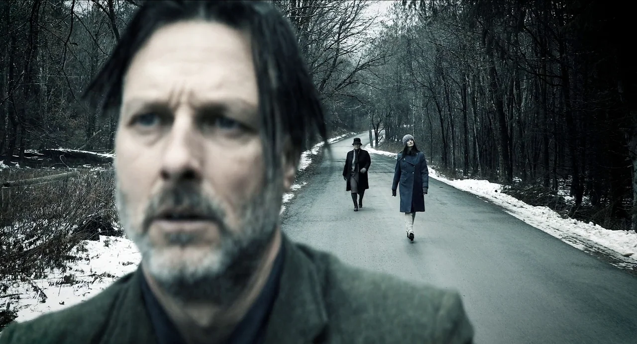 Where The Devil Roams (2023) movie still: medium — A wet asphalt road stretches through a dense, leafless forest dusted with patches of snow. One…