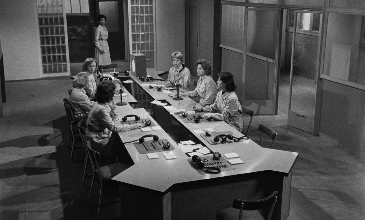 Witness In The City (1959) movie still: wide — A spacious 1960s communication center featuring a long, angular desk with multiple telephone…