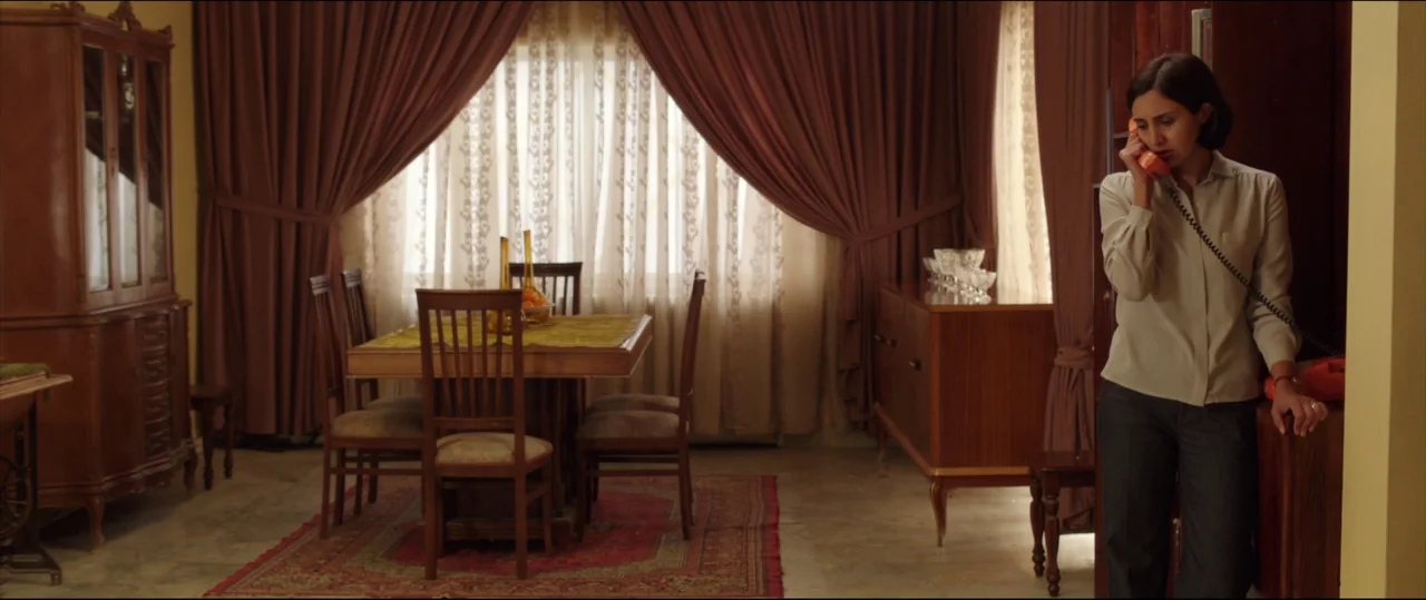 Under The Shadow (2016) movie still: medium — A formal dining room features heavy brown drapes and a wooden dining set. A woman in a beige shirt…