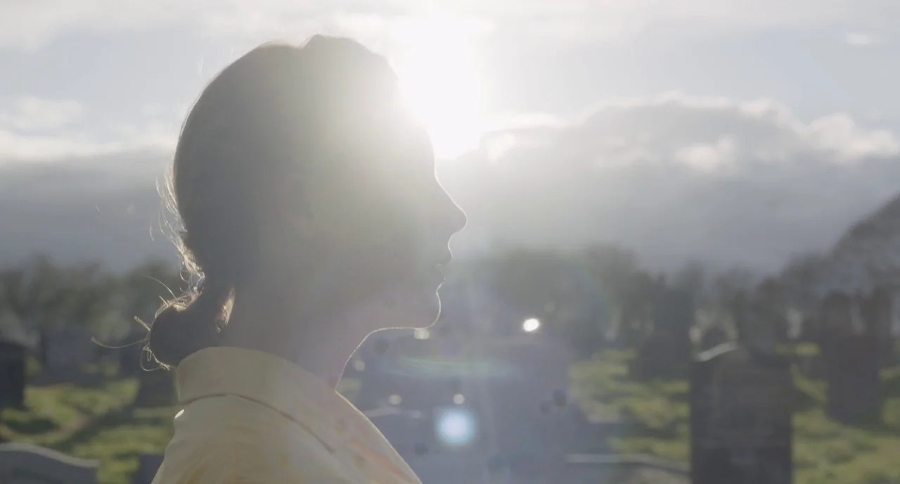 Brooklyn (2015) movie still: closeup — A cemetery with various gravestones sits under a bright, overcast sky during the day. A woman in a yellow…