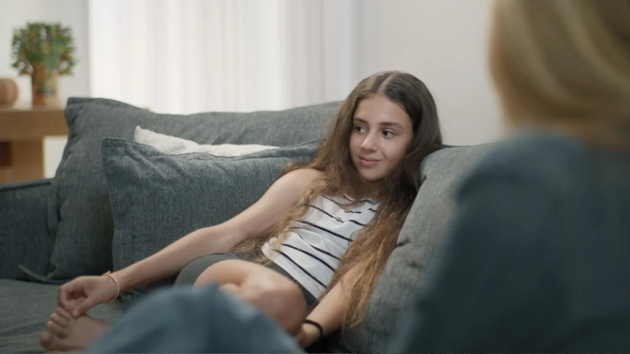 The Children of October 7 (2025) movie still: medium — A light-filled living room features a grey couch with soft throw pillows. A young girl sits…