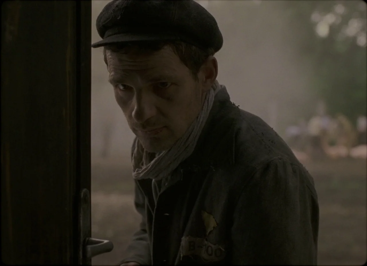 Son of Saul (2015) movie still: medium — An outdoor area features a wooden door frame partially obscuring the view. A gaunt man in a uniform jacket…