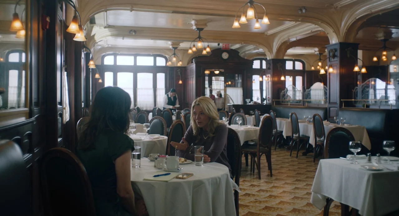 She Said (2022) movie still: medium — A restaurant dining room features dark wood paneling, ornate moldings, and white-clothed tables. Two women sit…