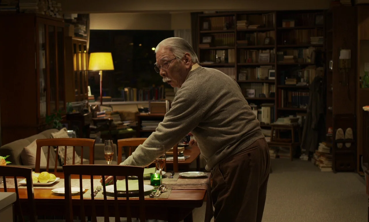 Like Someone In Love (2012) movie still: medium — A dimly lit room contains a dining table and wooden bookshelves full of books. An elderly man…