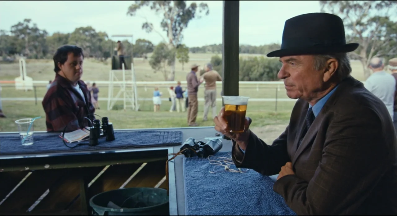 Ride Like a Girl (2019) movie still: medium — An outdoor bar at a racetrack features two men sitting at a table. The older man holds a plastic cup…