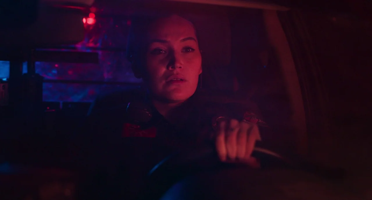 Eureka (2024) movie still: closeup — Interior of a police car at night illuminated by emergency lights. A female officer in uniform is driving and…