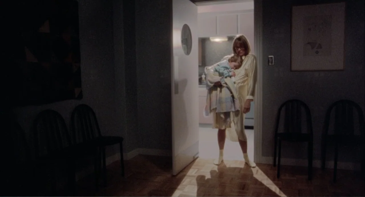 Baby Boom (1987) movie still: fullbody — A dark hallway with wooden floors connects to a brightly lit room through a door. A woman stands in the…