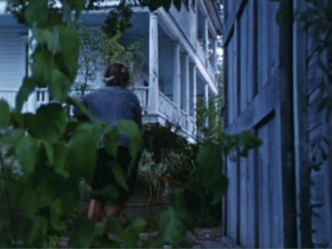It's Alive (1974) movie still: fullbody — A residential yard with a large white house and a wooden fence visible. A person in dark clothing walks…