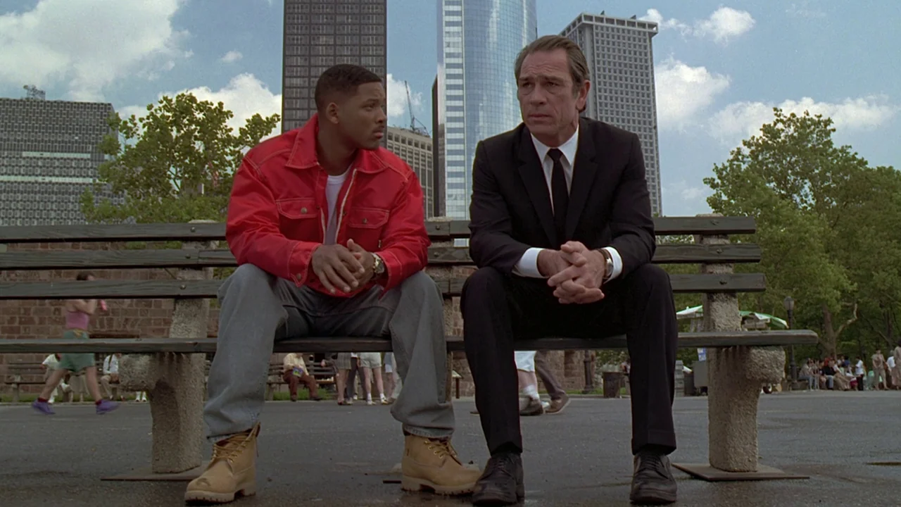 Men In Black (1997) movie still: fullbody — A public park with skyscrapers in the distant background under a bright cloudy sky. Two men sit on a…