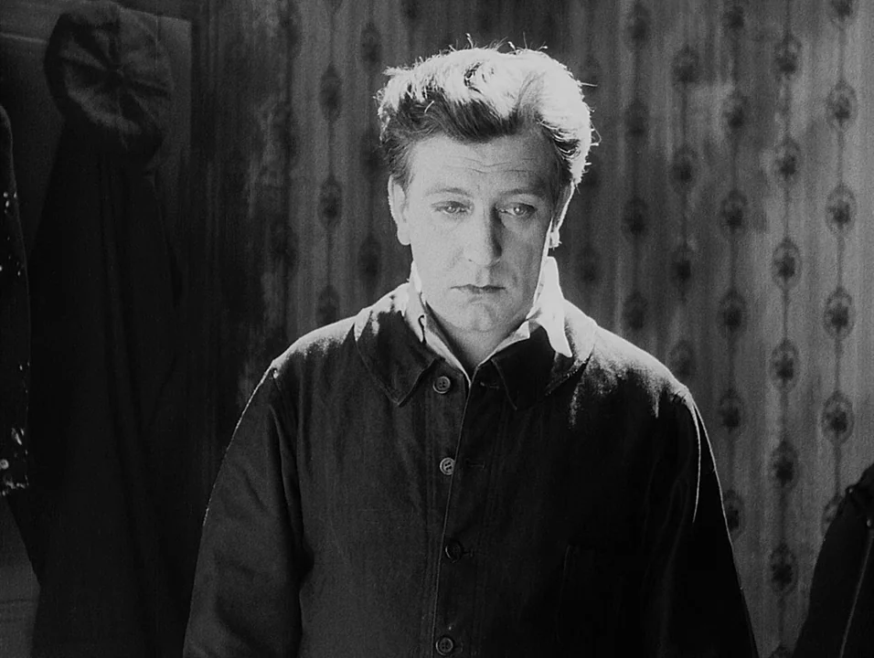 Coeur Fidele (1923) movie still: medium — A man stands indoors against a patterned wallpaper background. He stares forward in a static medium shot.