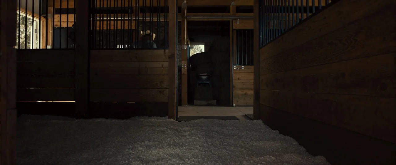 Frontera (2014) movie still: medium — A wooden horse stall with vertical railings features a person standing in the back. The stall is filled with…