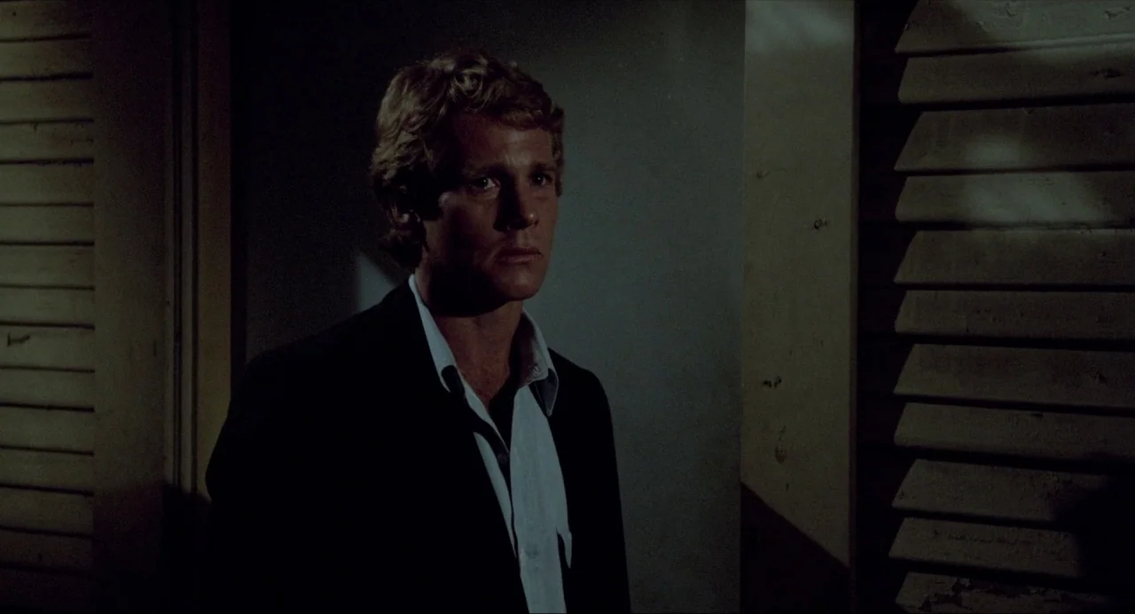 The Driver (1978) movie still: medium — A dimly lit interior space with wooden louvered shutters on both sides. A man in a dark blazer stands…
