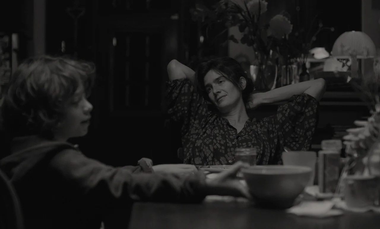 C’mon C’mon (2021) movie still: medium — A dimly lit room features a woman sitting at a table with a boy. The woman sits with her hands behind her…