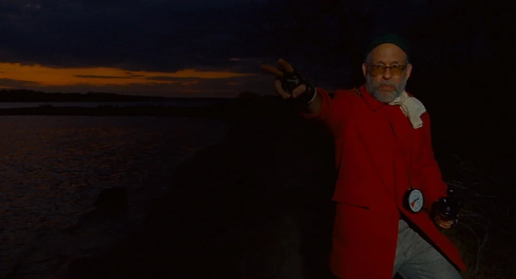 Moonrise Kingdom (2012) movie still: medium — A dark riverbank at twilight features a narrow band of orange light on the horizon. An elderly man in…