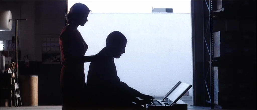 Punch Drunk Love (2002) movie still: medium — An interior space with stacked boxes and industrial equipment lit by a large door. A man sits while a…