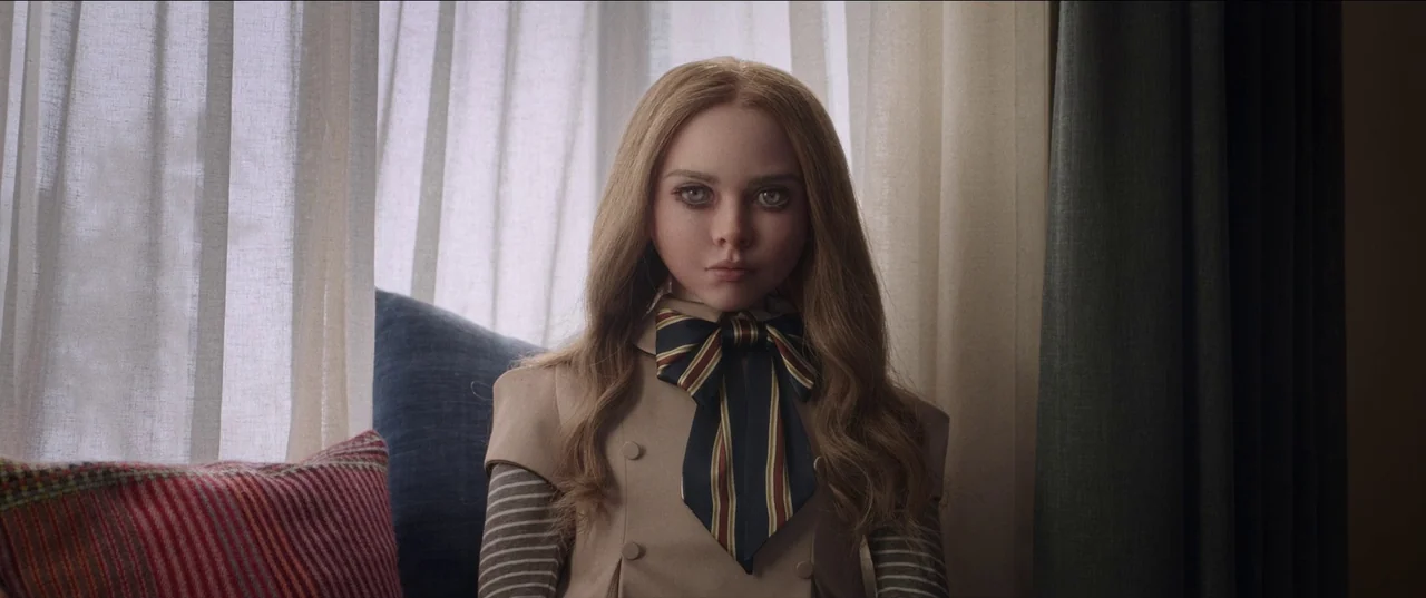 M3gan (2022) movie still: medium — A doll sits perfectly centered against a window covered by sheer white curtains. The doll has long hair and wears…