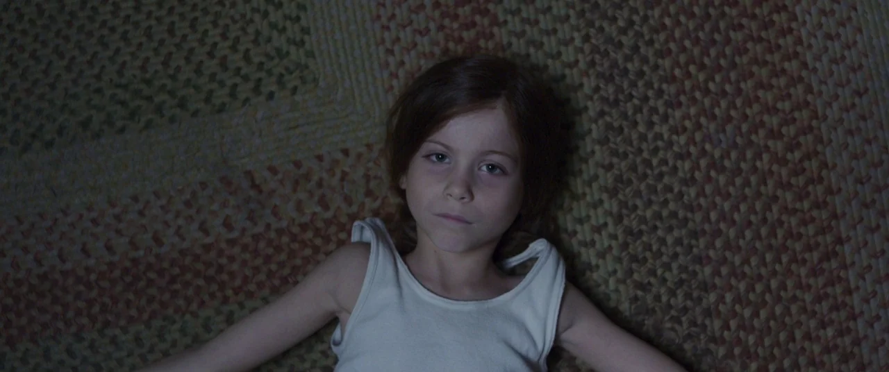 Room (2015) movie still: medium — A patterned woven rug in shades of red, beige and brown fills the frame under dim illumination. A young girl lies…