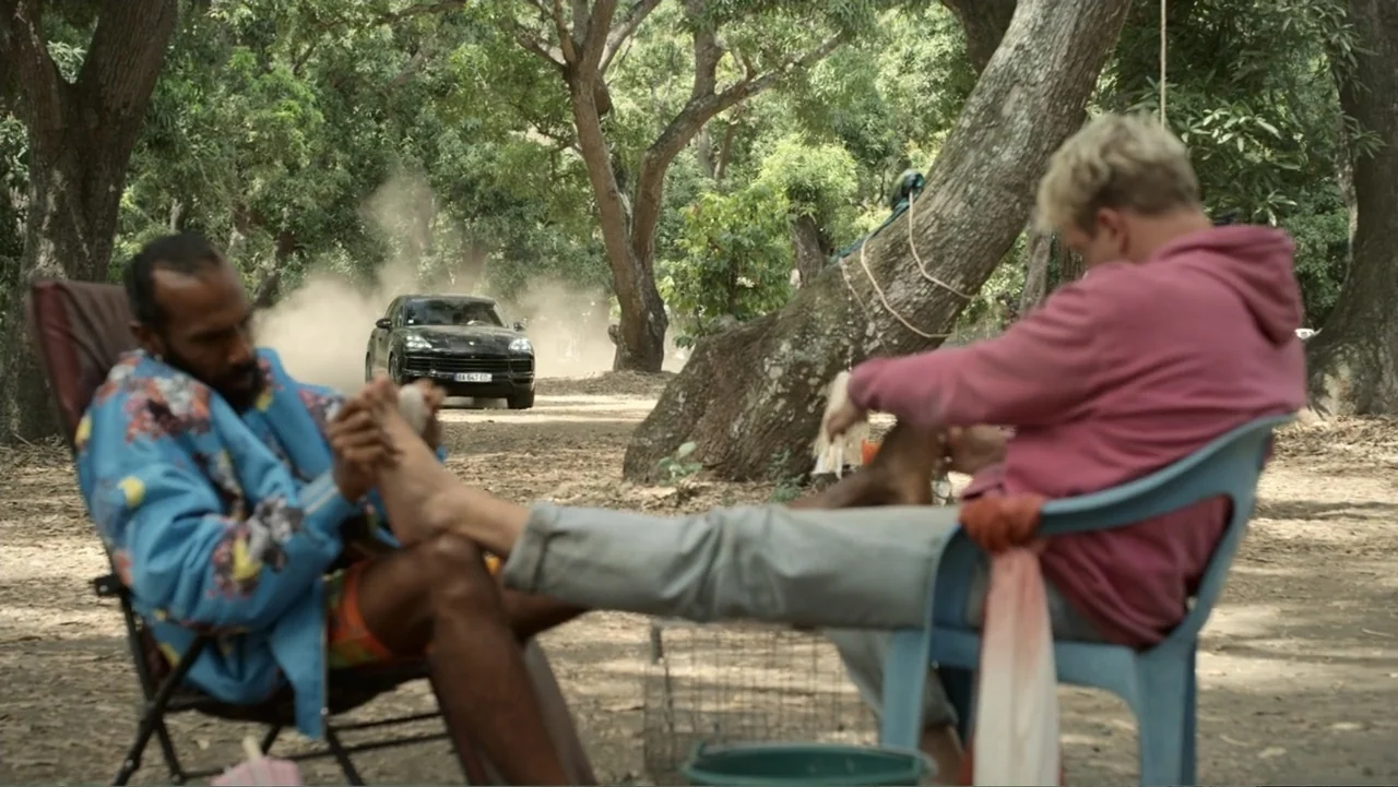 Unknown #1 movie still: fullbody — A dusty patch of ground is situated beneath a canopy of large trees. Two men sit in chairs facing each other…
