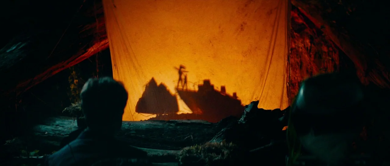 Swiss Army Man (2016) movie still: medium — A dark cave interior contains a glowing yellow cloth screen used for puppet projections. Two people sit…