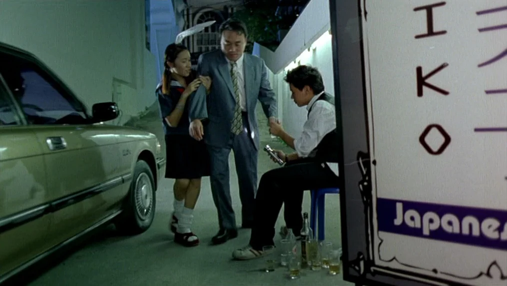 Last Life in the Universe (2003) movie still: wide — A night time curbside setting with a parked sedan and a Japanese signage board. A man in a suit…