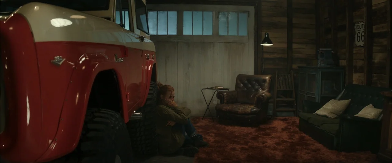 Maintenance Required (2025) movie still: wide — A dark wooden garage features a vintage truck, a leather armchair, and a shag rug. A woman sits on…