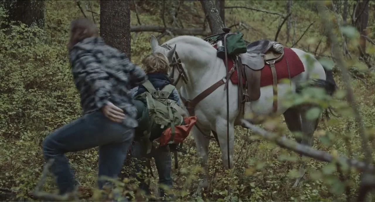 Cowboys (2014) movie still: medium — A dense patch of wilderness contains thick undergrowth and tall pine tree trunks. Two figures walk away from…