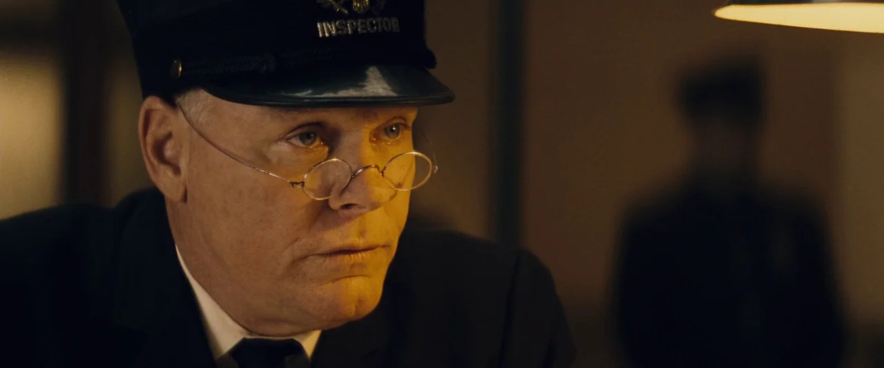 The Immigrant (2013) movie still: closeup — An older man in a uniform sits inside a dimly lit office space. He wears wire-rimmed spectacles and an…
