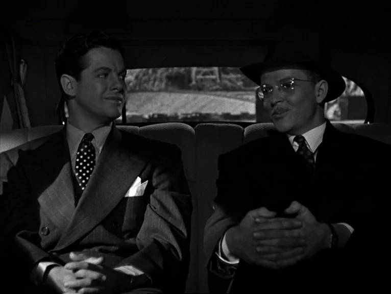 Saboteur (1942) movie still: medium — Two men sit together in the back seat of a moving vehicle. They face toward each other in conversation while…