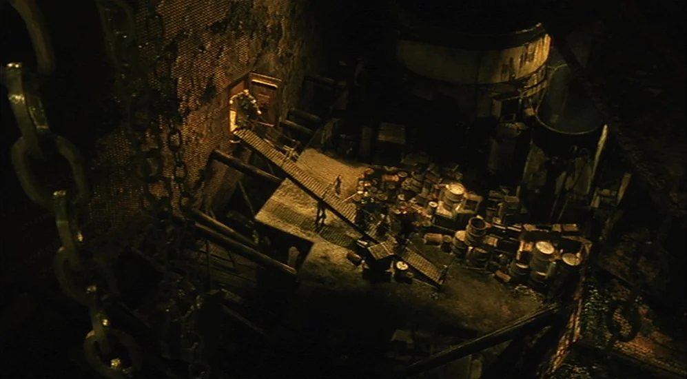 The City Of Lost Children movie still: establishing — A dark, cramped industrial basement features metal ramps and stacks of debris. Massive iron…