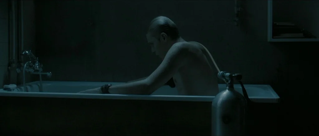 Lasting (Nieulotne) (2013) movie still: medium — A dimly lit room contains a white bathtub with dark wall panels. A young man sits in the tub with a…