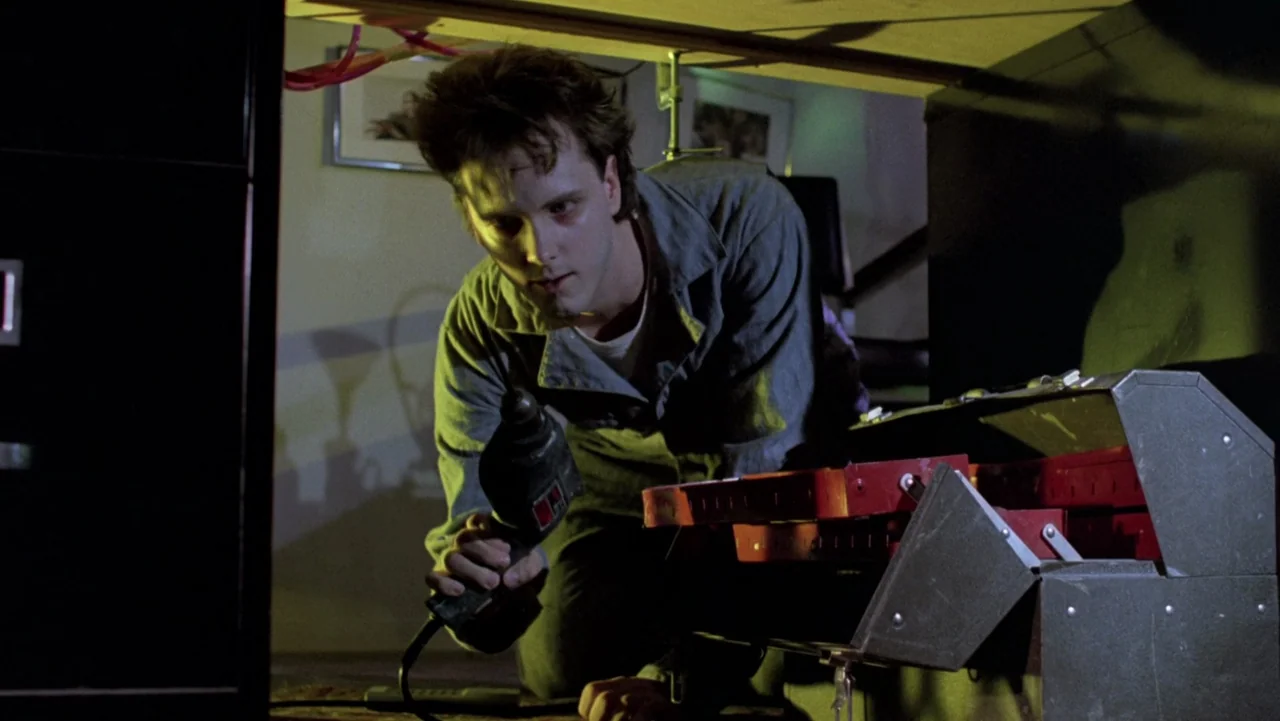 Frankenhooker (1990) movie still: medium — A dark, cramped space filled with mechanical equipment and a small metal desk. The man kneels on the…