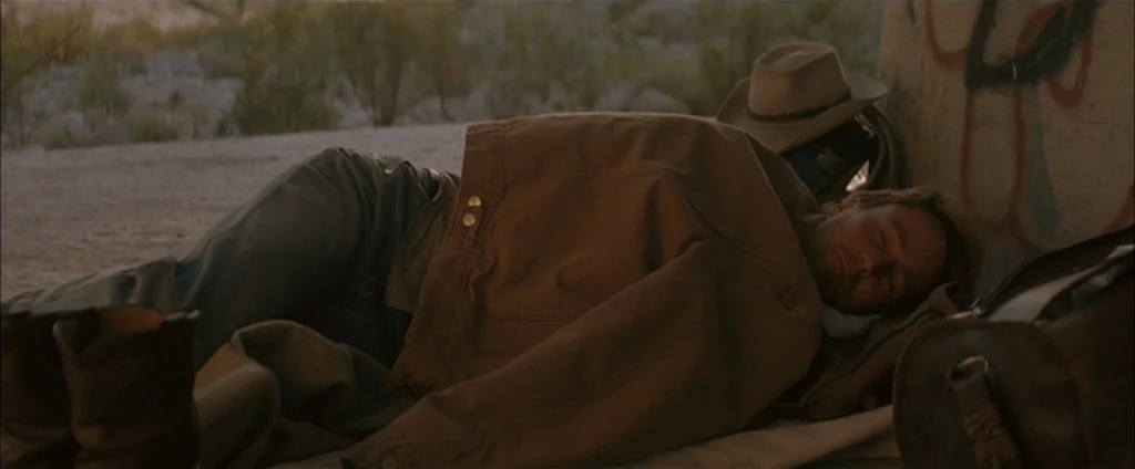 Down In The Valley (2005) movie still: medium — A man sleeps on a blanket beneath a concrete bridge structure with graffiti markings. The man rests…