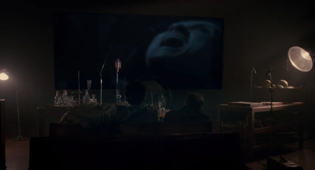 Berberian Sound Studio (2012) movie still: wide — A dark, dimly lit room contains a large projection screen displaying a person's face. Two men are…