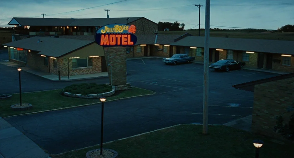 A Serious Man (2009) movie still: establishing — A single-story U-shaped motel exterior at dusk features a prominent lit neon sign. Two vintage cars…