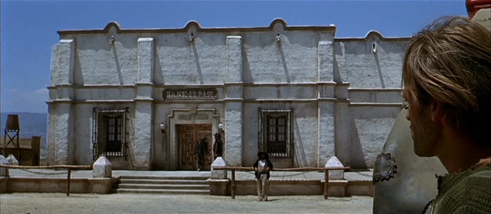 For A Few Dollars More (1965) movie still: wide — A stucco building labeled Bank El Paso stands in a dry, sunlit dusty plaza. A man in the…